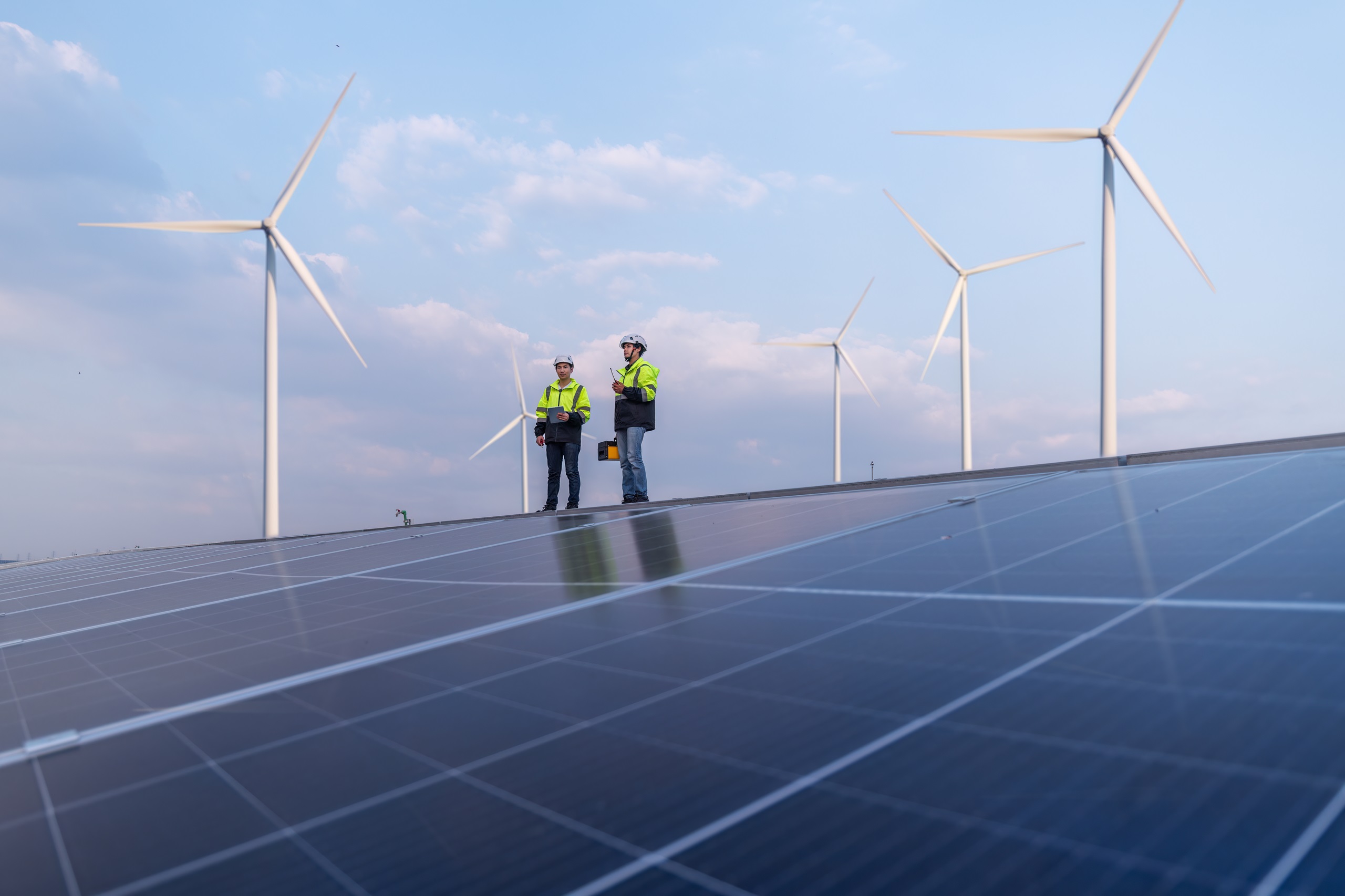 Zwei TÜV NORD-Ingenieure prüfen Solarmodule vor Windrädern unter blauem Himmel