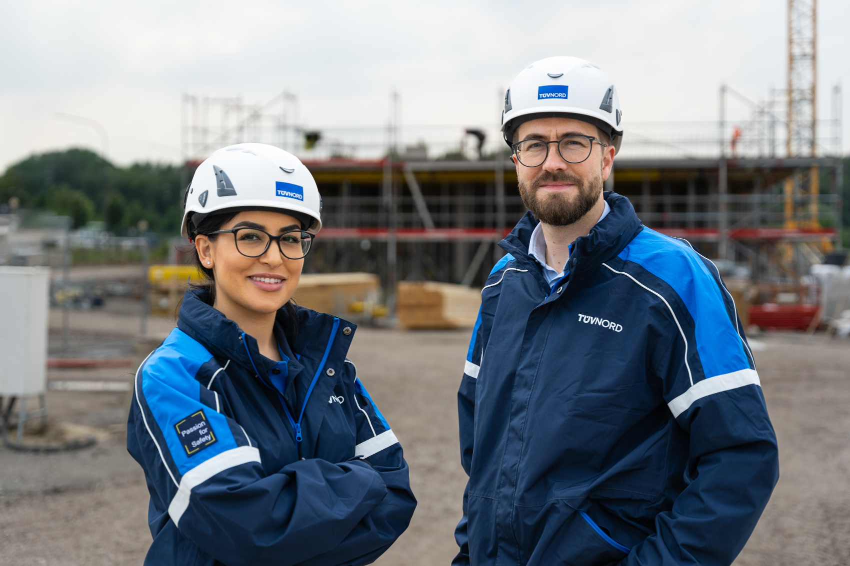 Zwei BauingenieurInnen mit Schutzhelmen und blauer Arbeitskleidung stehen vor einer Baustelle und lächeln in die Kamera.
