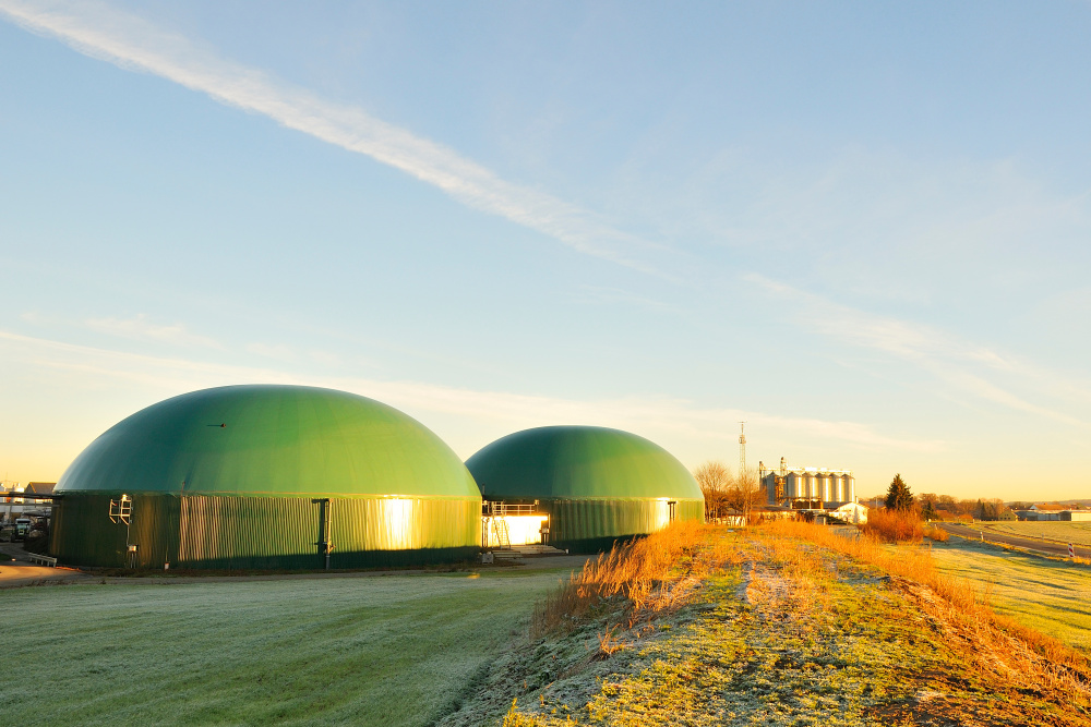 Zwei grüne Kuppelbauten einer Biogasanlage in ländlicher Umgebung bei Sonnenaufgang.