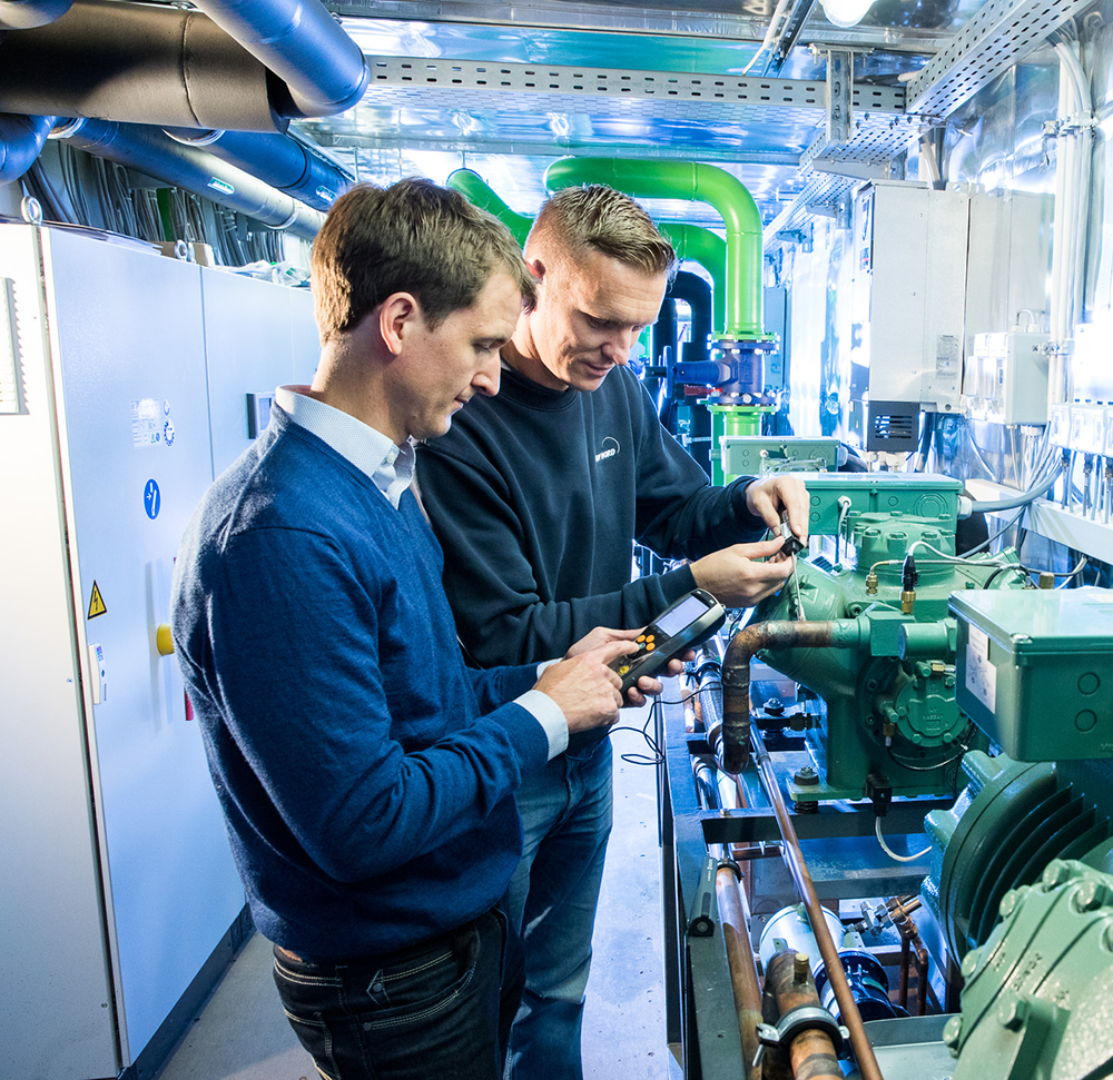 Zwei Mitarbeitende stehen im Prüfbereich der Kälte-, Klima- und Lüftungstechnik bei TÜV NORD und blicken auf technische Geräte, die sie in den Händen halten.