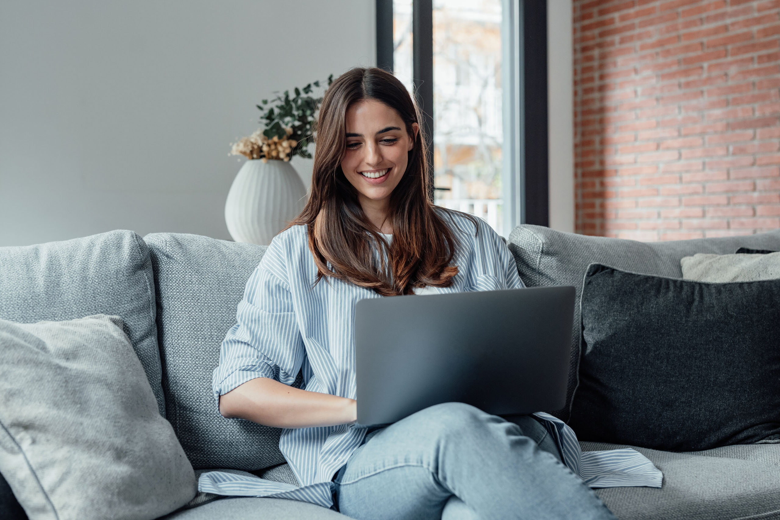 Junge Frau sitzt auf einem Sofa und arbeitet lächelnd an einem Laptop