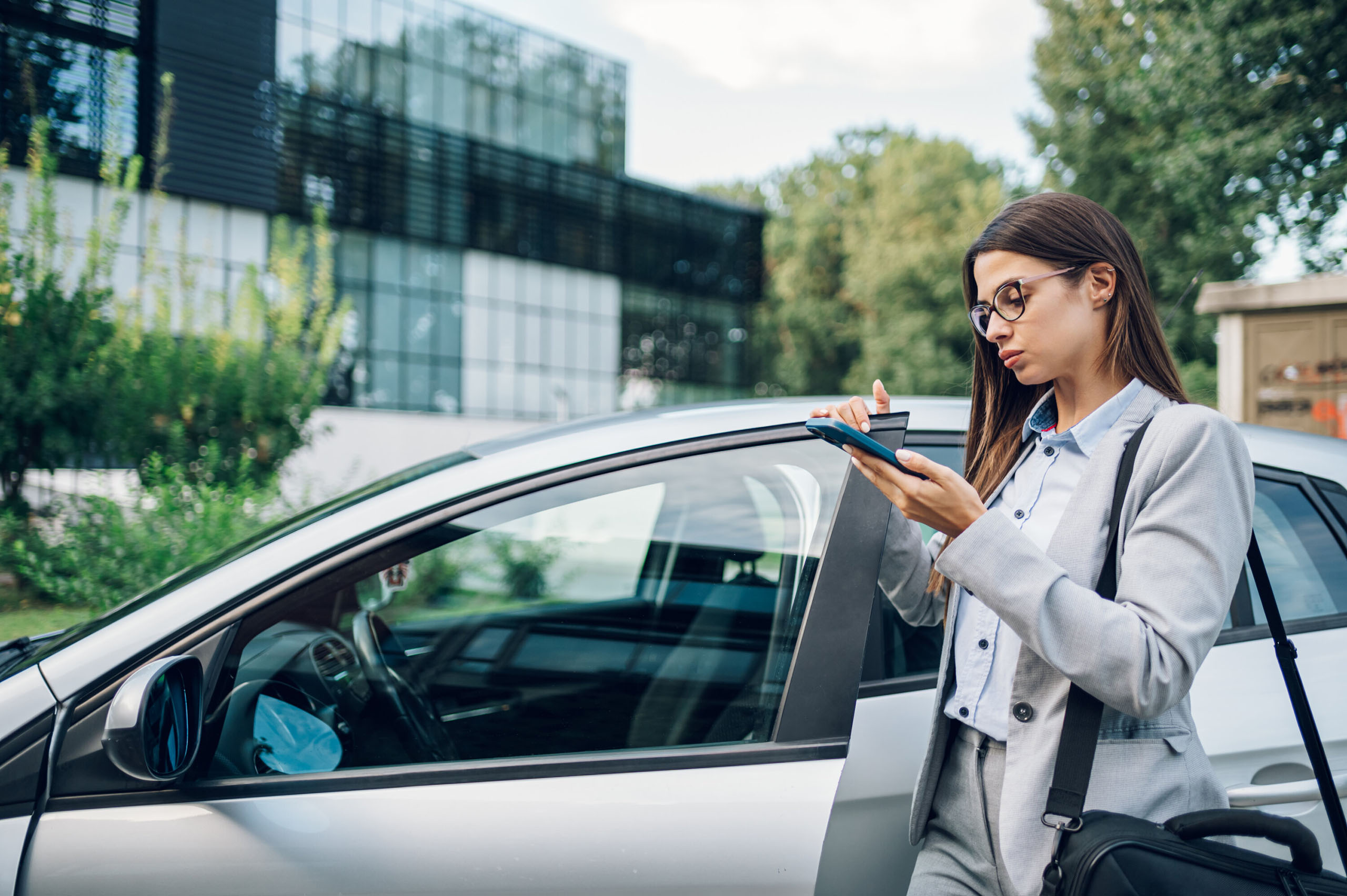 Frau im Business-Anzug mit Brille steht neben einem Auto und notiert auf einem Tablet