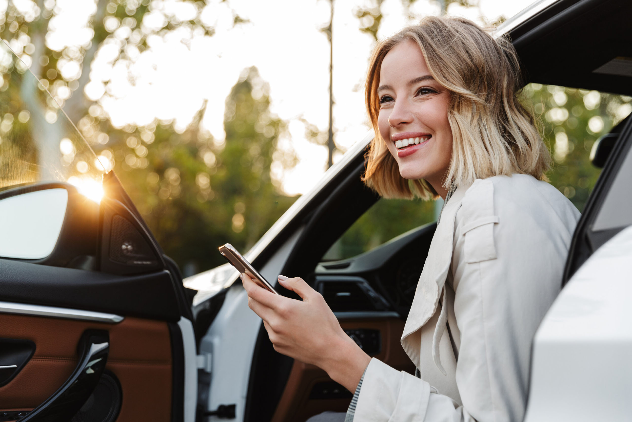 Lächelnde junge Frau sitzt mit Smartphone in der Hand in der offenen Tür eines Autos