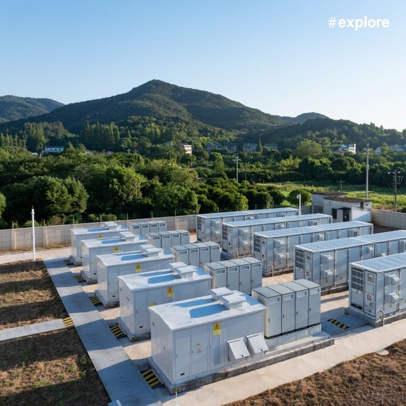 Eine moderne Energieanlage mit mehreren weißen Containergebäuden, die in einer grünen Landschaft mit Hügeln im Hintergrund liegt. Der Himmel ist klar und blau, und der Hashtag "#explore" ist oben rechts im Bild zu sehen.