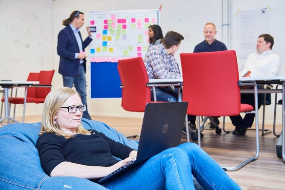 Ein Arbeitsraum mit mehreren Personen und roten Stühlen. Im Vordergrund sitzt eine Person mit Brille auf einem blauen Sitzsack mit Laptop, während im Hintergrund andere an einer Pinnwand arbeiten.