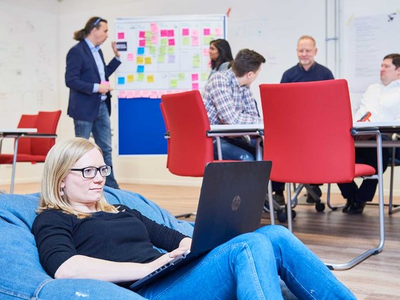 Ein Arbeitsraum mit mehreren Personen und roten Stühlen. Im Vordergrund sitzt eine Person mit Brille auf einem blauen Sitzsack mit Laptop, während im Hintergrund andere an einer Pinnwand arbeiten.