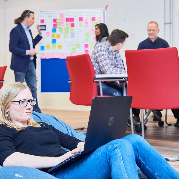 Ein Arbeitsraum mit mehreren Personen und roten Stühlen. Im Vordergrund sitzt eine Person mit Brille auf einem blauen Sitzsack mit Laptop, während im Hintergrund andere an einer Pinnwand arbeiten.
