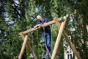 Der Arbeiter steht auf einer Leiter und arbeitet an der Montage einer Holzkonstruktion mit Ketten. Das Spielgerät befindet sich in einem Park mit grünen Bäumen im Hintergrund.