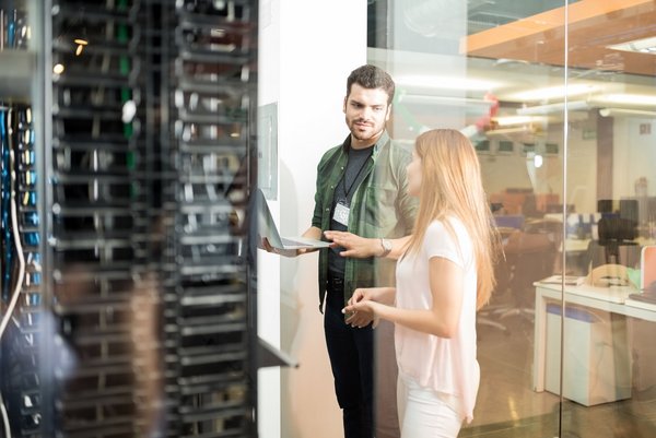 Zwei IT-Fachkräfte stehen im Serverraum und besprechen Daten auf einem Laptop vor einem Serverschrank