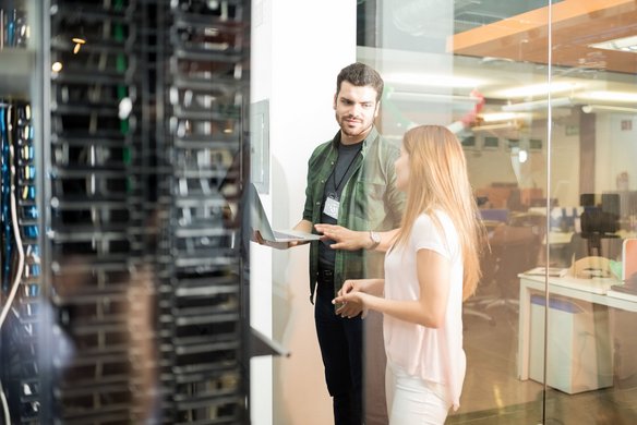 Zwei IT-Fachkräfte stehen im Serverraum und besprechen Daten auf einem Laptop vor einem Serverschrank