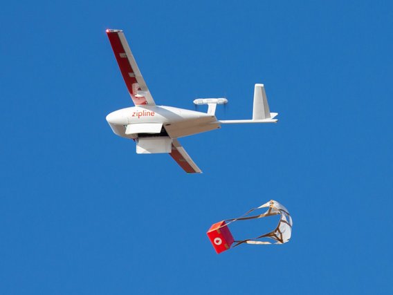 Eine weiße Zipline-Drohne mit roten Markierungen am Heck fliegt vor blauem Himmel und liefert ein rotes Paket mit Fallschirm ab