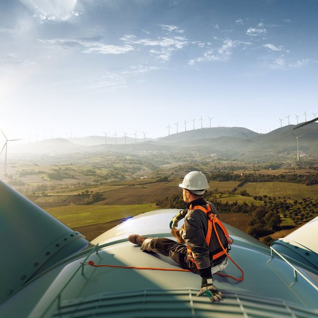 Techniker mit Sicherheitsausrüstung sitzt auf einer Windkraftanlage mit Blick auf ein Tal und weitere Windräder.