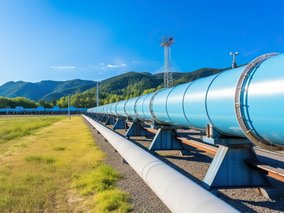 Große silberne Pipeline verläuft über grüne Wiesen vor bewaldeten Bergen unter blauem Himmel mit weißen Wolken.
