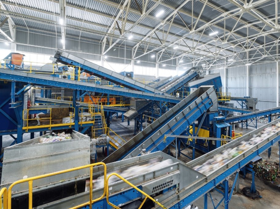 Moderne Recycling- oder Abfallsortieranlage mit blauen Förderbandsystemen in einer weitläufigen Industriehalle mit Metallkonstruktion und heller Beleuchtung.