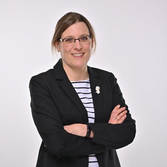 Britta Schacht mit Brille in einem schwarzen Blazer über einem weiß-schwarz gestreiften Shirt. Sie lächelt in die Kamera und steht mit verschränkten Armen vor einem neutralen grauen Hintergrund.