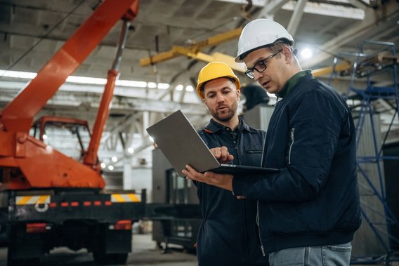 Zwei Ingenieure mit Schutzhelmen prüfen Produktionsdaten auf einem Laptop in der Werkshalle