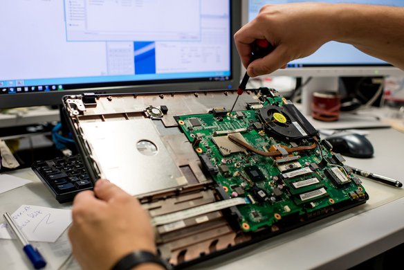 Eine Person repariert einen Laptop und arbeitet mit einem Schraubenzieher am offenen Motherboard, während ein Computer-Bildschirm im Hintergrund zu sehen ist.