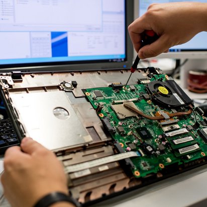 Eine Person repariert einen Laptop und arbeitet mit einem Schraubenzieher am offenen Motherboard, während ein Computer-Bildschirm im Hintergrund zu sehen ist.
