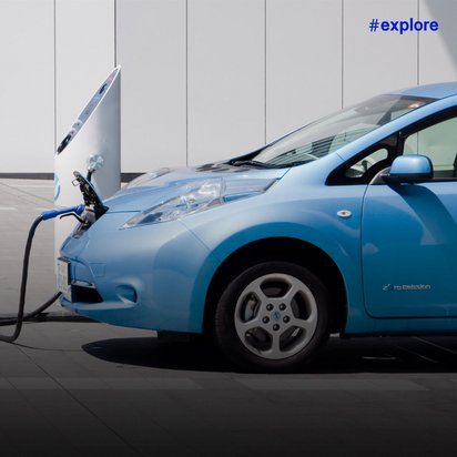 Ein blauer Nissan Leaf Elektroauto wird an einer Ladestation vor einem modernen Gebäude aufgeladen. Das Fahrzeug trägt den "Zero Emission" Schriftzug und zeigt die geöffnete Motorhaube während des Ladevorgangs.