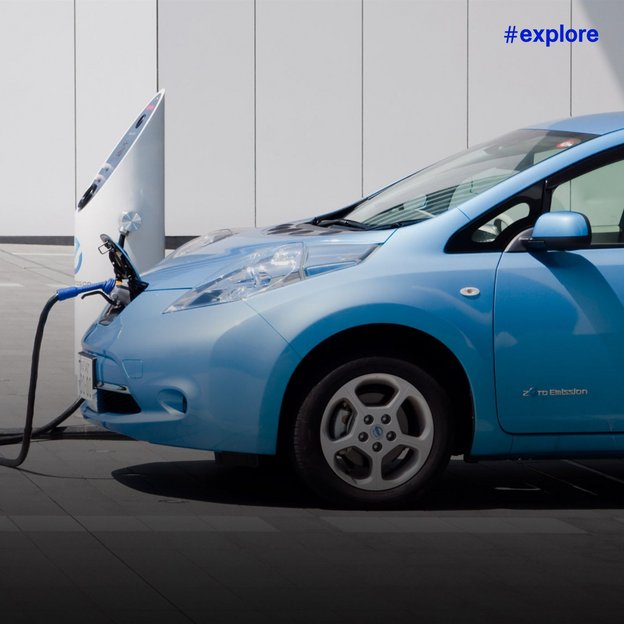 Ein blauer Nissan Leaf Elektroauto wird an einer Ladestation vor einem modernen Gebäude aufgeladen. Das Fahrzeug trägt den "Zero Emission" Schriftzug und zeigt die geöffnete Motorhaube während des Ladevorgangs.