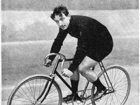 Ein Radfahrer (Ludovic Morin) in schwarzer Kleidung auf einem historischen Rennrad aus der Frühzeit des Radsports. Schwarzweißfoto zeigt einen sportlichen Fahrer in dynamischer Haltung.