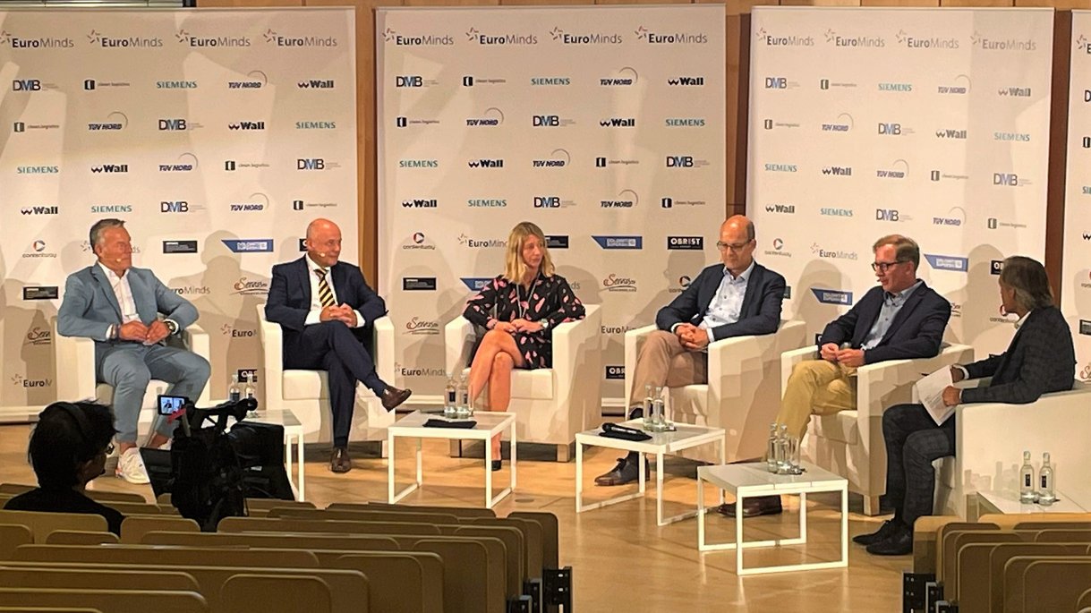 Eine Podiumsdiskussion mit mehreren Teilnehmern auf weißen Stühlen auf einer Bühne mit EuroMinds-Branding im Hintergrund.