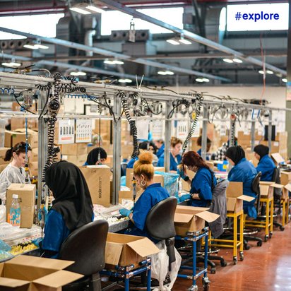 Blick in eine Produktionshalle mit Mitarbeitern in blauen Arbeitskleidungen. Sie sitzen an Arbeitsstationen mit Kartons und verstellbaren Stühlen, ausgestattet mit beweglichen Kabeln und Halterungen. Die Halle hat eine hohe Decke mit Industriebeleuchtung.WiederholenClaude kann Fehler machen. Bitte überprüfen Sie die Antworten.