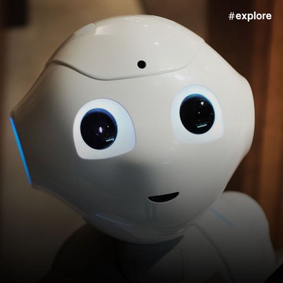 Ein weißer humanoider Roboter mit großen, runden schwarzen Augen und einem kleinen Mund blickt direkt in die Kamera. Im Hintergrund sind warme Holztöne und eine Backsteinwand zu sehen, die eine gemütliche Innenraumatmosphäre schaffen.