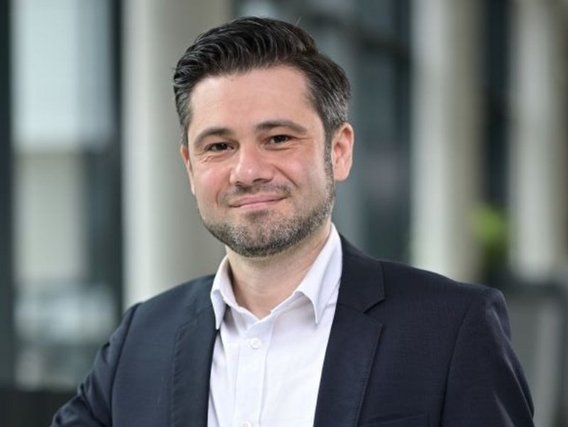 Porträtaufnahme eines lächelnden Mannes mit dunklen, zurückgekämmten Haaren und leichtem Bart in dunklem Anzug und weißem Hemd vor einem unscharfen Bürogebäude-Hintergrund.