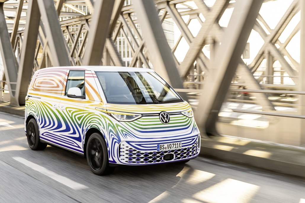 Ein weißer Volkswagen-Van mit wellenförmigen bunten Streifen fährt über eine moderne Stahlbrücke in urbaner Umgebung. Das Elektrofahrzeug im futuristischen Design repräsentiert nachhaltige Mobilität in der Stadt.