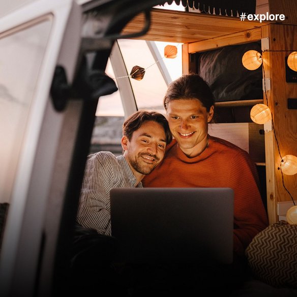 Zwei lächelnde Männer sitzen gemütlich mit einem Laptop in einem beleuchteten Wohnmobil bei Abenddämmerung. Die warme Atmosphäre mit Lichterketten zeigt das entspannte Leben auf Reisen mit moderner Technologie.