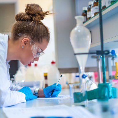 Frau im Labor trägt Schutzbrille und Handschuhe und schreibt Ergebnisse auf