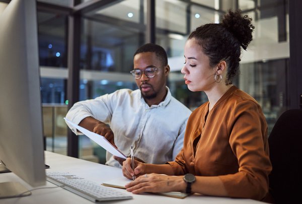 Zwei Kollegen arbeiten abends gemeinsam am Computer und besprechen Unterlagen im modernen Büro