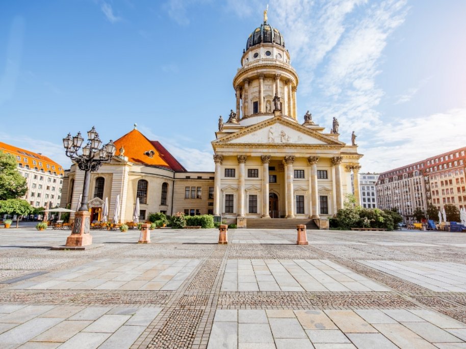 Ein historisches Gebäude mit Säulen und Kuppel steht auf einem gepflasterten Platz umgeben von Bäumen und Häusern.