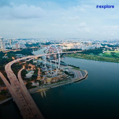 Luftaufnahme einer Küstenstadt mit Hochhäusern, einem großen Riesenrad am Ufer, geschwungenen Autobahnbrücken und einem weiten Gewässer im Hintergrund.