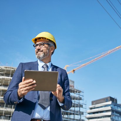 Ein Mann in Anzug und Schutzhelm steht vor einer Baustelle und nutzt ein Tablet. Im Hintergrund sind Rohbauten und ein Baukran zu sehen. Baustellenmanagement mit digitaler Unterstützung