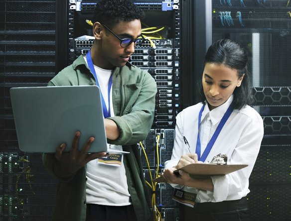 Zwei IT-Fachkräfte prüfen mit Laptop und Klemmbrett Server in einem Rechenzentrum.