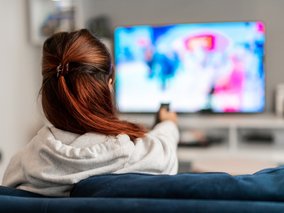 Rückansicht einer Person mit rotbraunem Haar in einem grauen Pullover, die auf einem Sofa sitzt und mit einer Fernbedienung fernsieht.