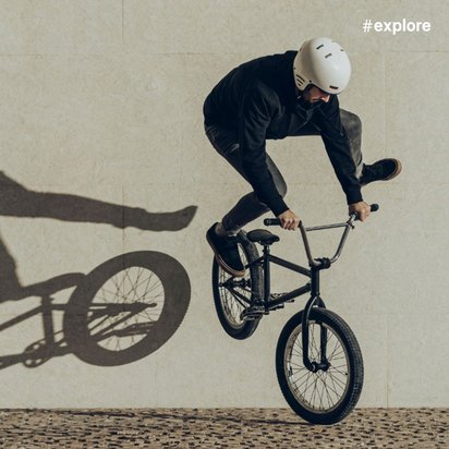 Ein BMX-Fahrer in schwarzer Kleidung und weißem Helm, der einen Trick auf einem schwarzen Fahrrad vor einer beigen Wand vollführt, mit einem dramatischen Schatten.
