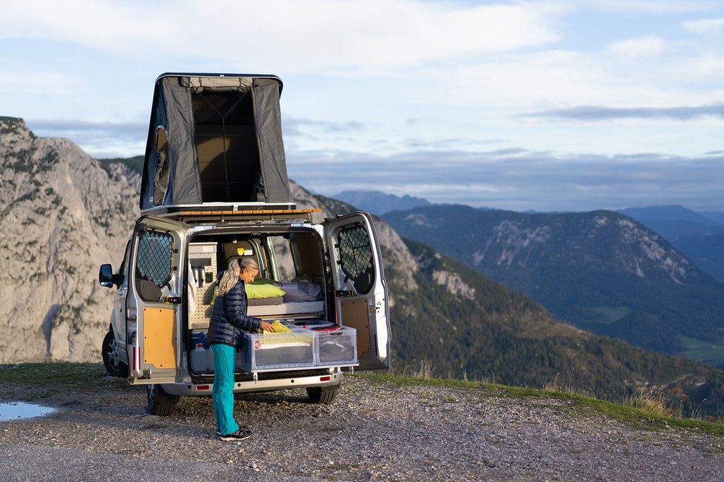 Eine Frau in türkisfarbener Hose und dunkler Jacke steht vor einem weißen Campingvan mit geöffneten Hecktüren und aufgeklapptem Dachzelt. Im Hintergrund erstreckt sich eine bergige Landschaft mit dramatischen Felsformationen und bewölktem Himmel.