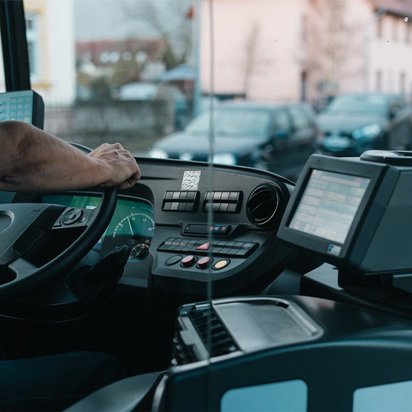 Ein Busfahrer steuert ein Fahrzeug durch eine städtische Straße. Das Cockpit zeigt moderne Bedienelemente und ein digitales Display.