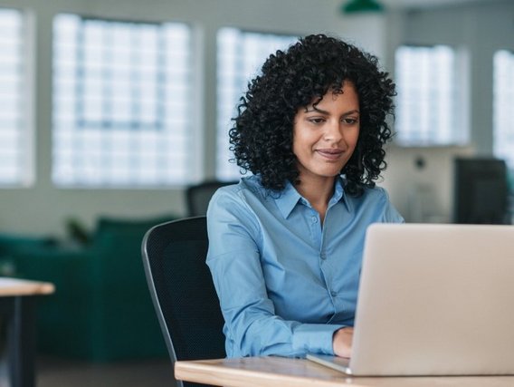 Frau mit lockigen schwarzen Haaren in hellblauem Hemd arbeitet lächelnd am Laptop in modernem Büro.