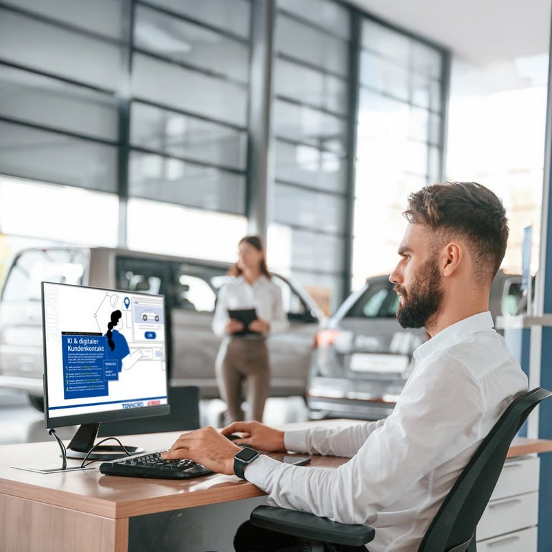 Mitarbeiter am Computer in einem Autohaus, Bildschirm zeigt Informationen über digitalen Kundenkontakt. Autos und Person mit Tablet im Hintergrund.