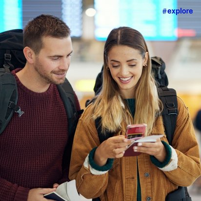Junges Reisepaar mit Rucksäcken schaut gemeinsam lächelnd auf ein Smartphone in einer modernen Flughafenhalle mit Anzeigetafeln im Hintergrund.