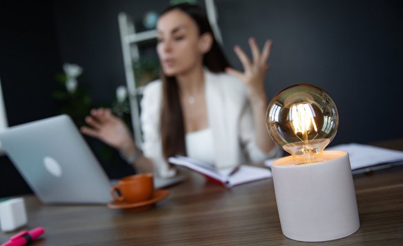 Frau im Gespräch vor Laptop mit Notizbuch, im Vordergrund eine leuchtende Glühbirne als Symbol für Ideen.