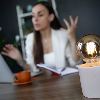 Frau im Gespräch vor Laptop mit Notizbuch, im Vordergrund eine leuchtende Glühbirne als Symbol für Ideen.