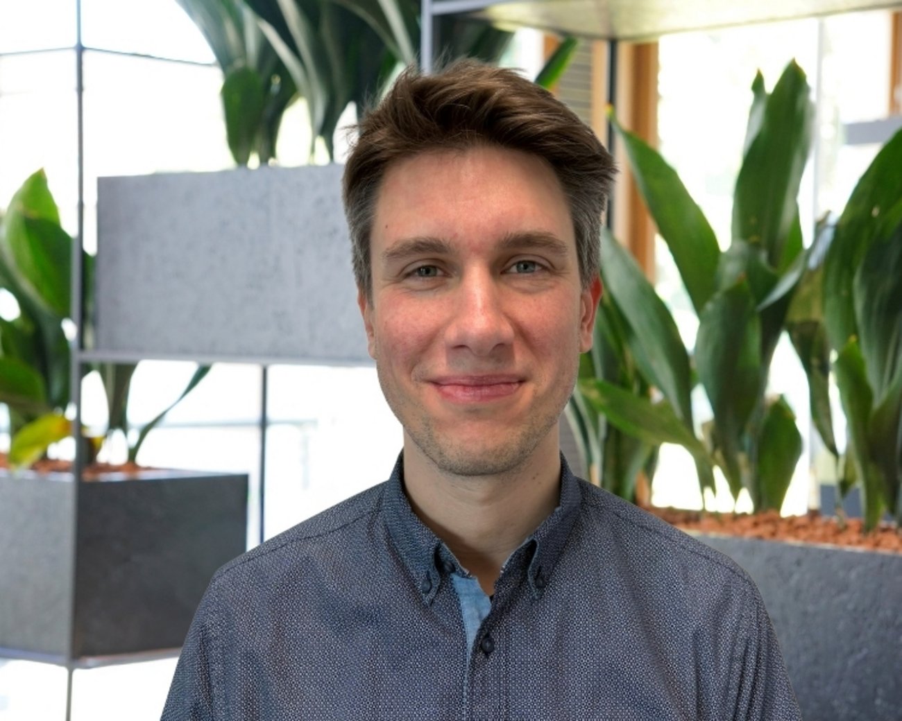 Foto von Niklas Skotorzick Person mit kurzem dunklem Haar trägt ein blaues Hemd mit kleinem Muster und steht vor mehreren rechteckigen Pflanzgefäßen mit großen grünen Zimmerpflanzen. Im Hintergrund sind helle Fensterflächen und ein metallisches Regalsystem sichtbar.