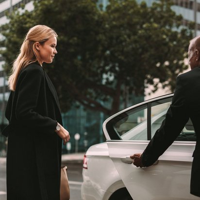 Frau steigt in ein weißes Auto ein, das von einem Fahrer an einer Stadtstraße geöffnet wird