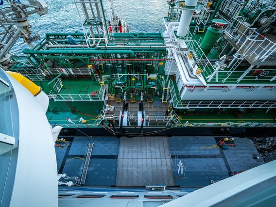 Vogelperspektive auf das grüne Arbeitsdeck eines Schiffes mit komplexer Rohrleitungsanlage, weißen Geländern und mehreren Arbeitern, umgeben von blauem Meerwasser.