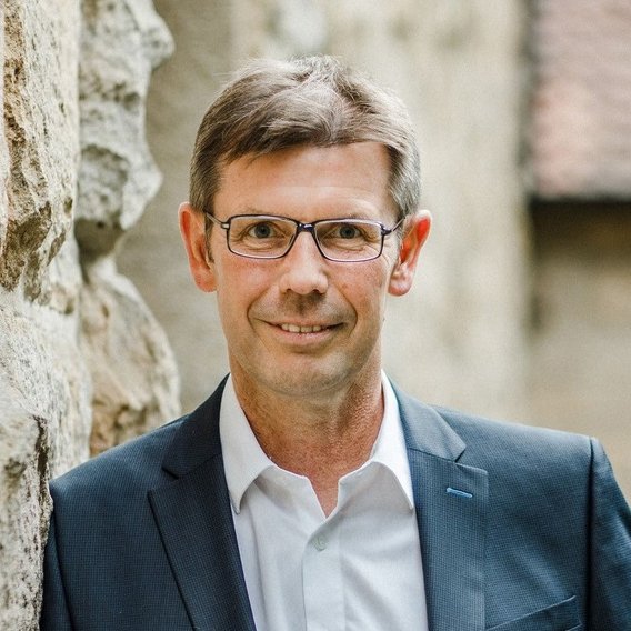 Ein Porträt von Gerd Bünker, der eine Brille trägt und ein weißes Hemd sowie einen grauen Anzug anhat.